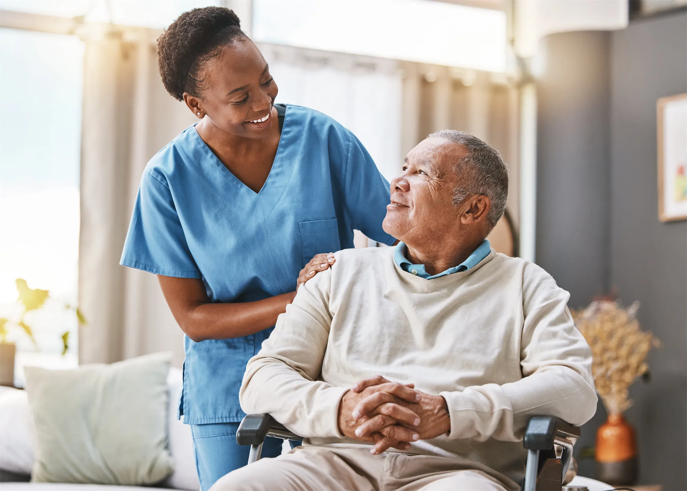 Photo of black woman in blue medical scrubs standing next to an older black man sitting in a wheel chair. Both are smiling at each other.